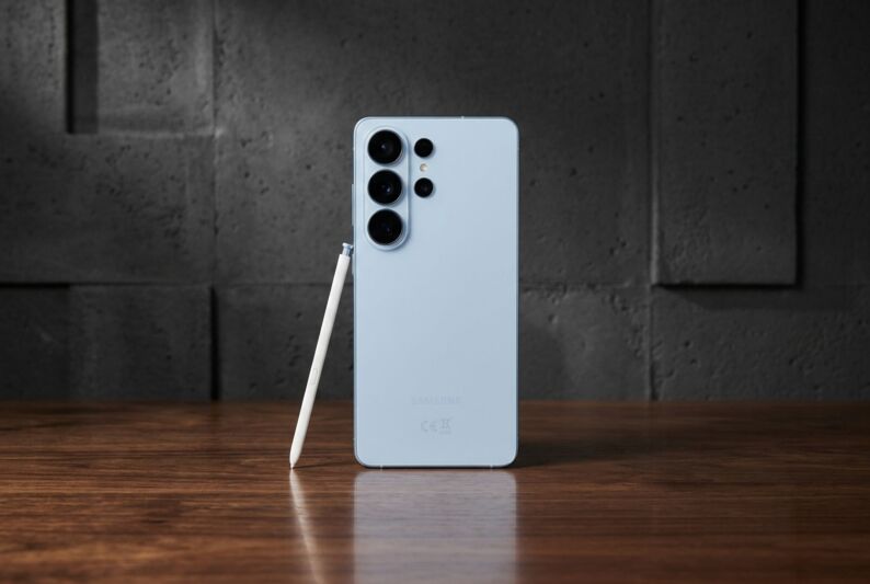 Smartphone Samsung bleu clair avec stylet posé sur une table en bois devant un mur gris.