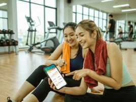 Deux femmes regardent une application de fitness sur un smartphone dans une salle de sport, éclairage naturel, photo.