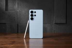 Smartphone Samsung bleu clair avec stylet posé sur une table en bois devant un mur gris.